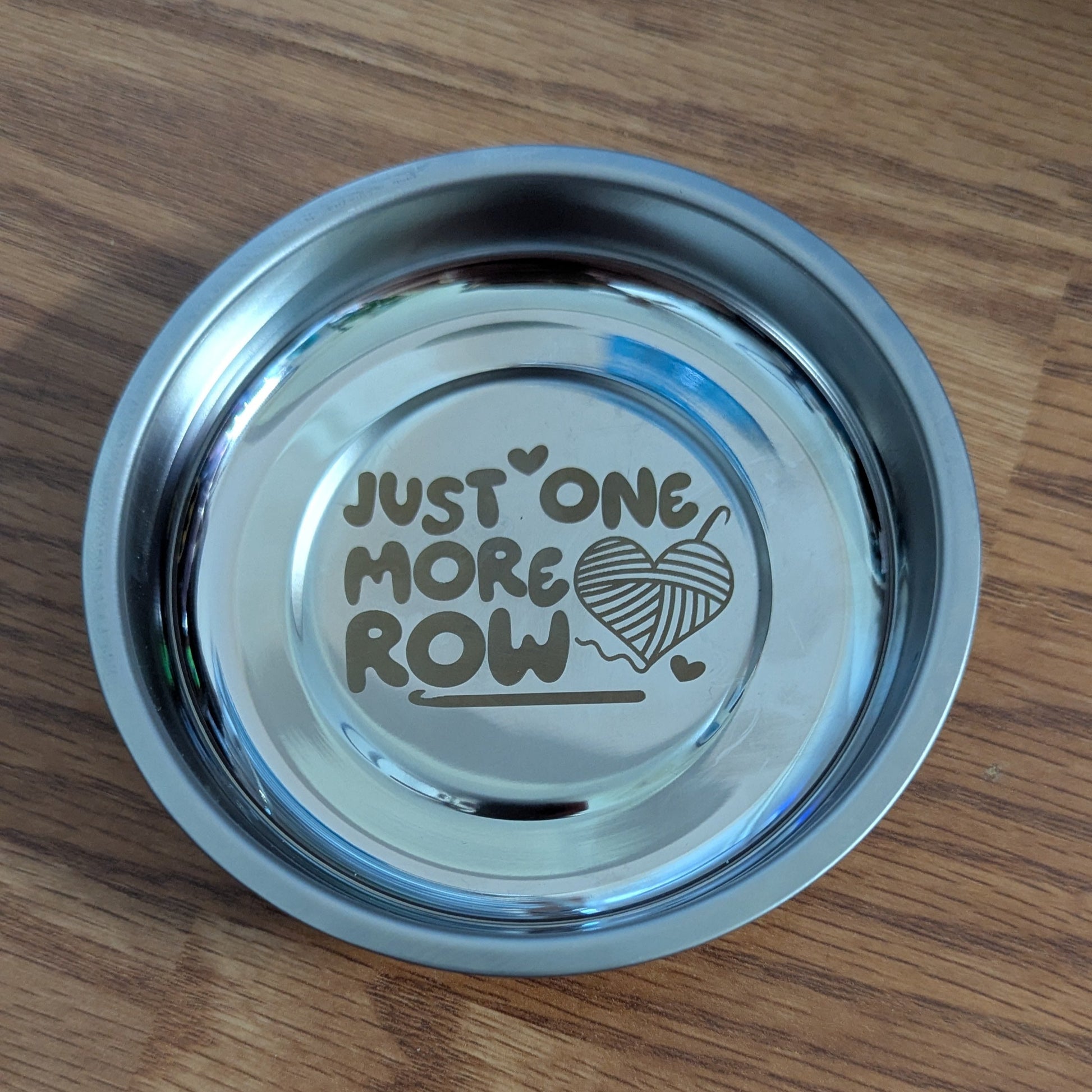 Metallic yarn bowl with 'Just One More Row' text on a wooden surface