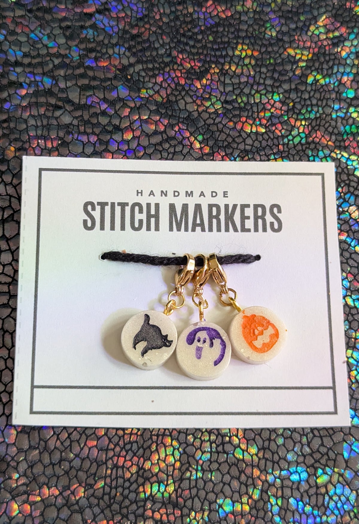 Three handmade circular stitch markers with cat, ghost, and pumpkin designs, attached to a black cord and displayed on a white card labeled 'HANDMADE STITCH MARKERS' against a holographic textured background.