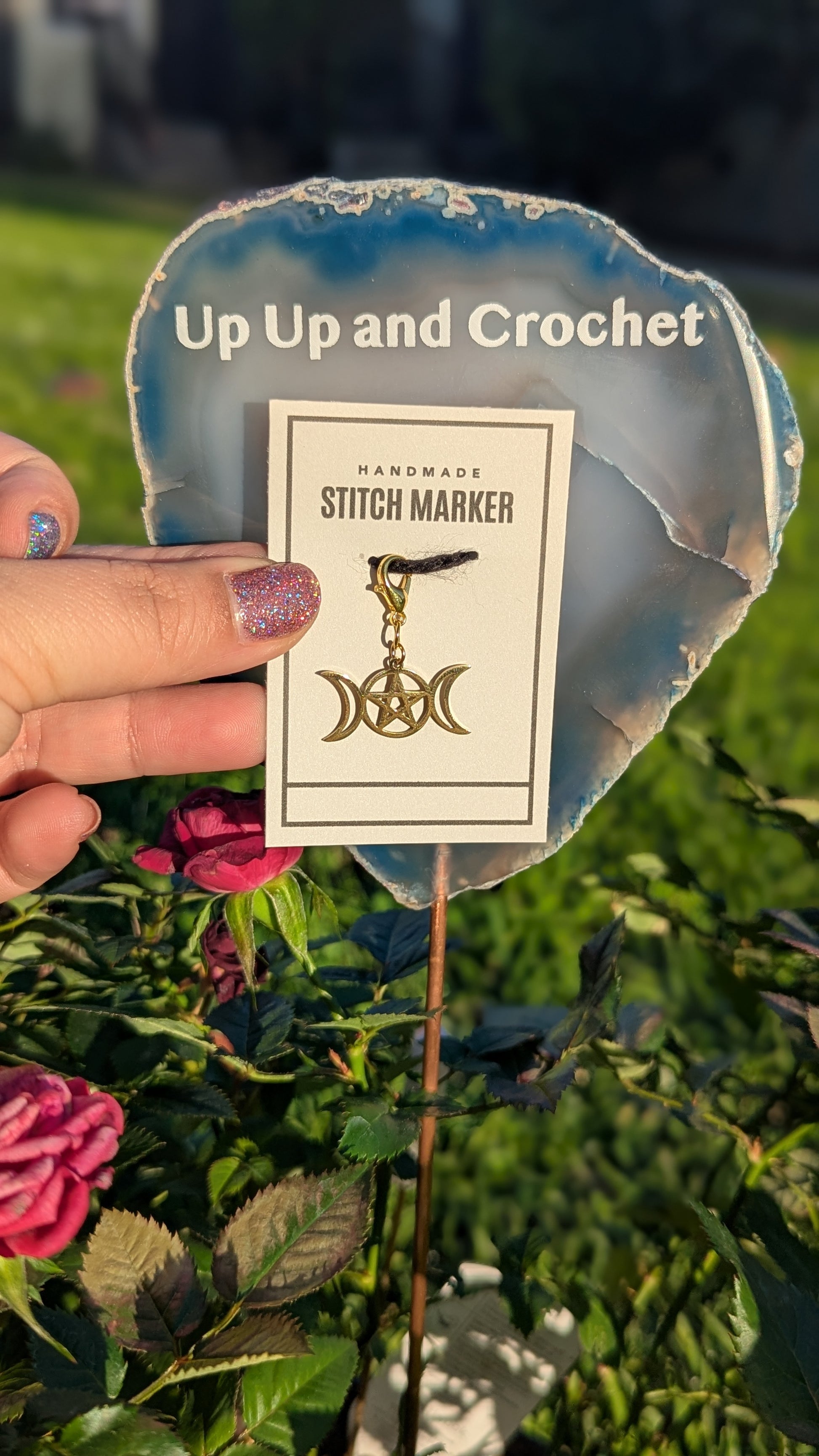 Handmade triple moon stitch marker with packaging held by a hand against a natural background